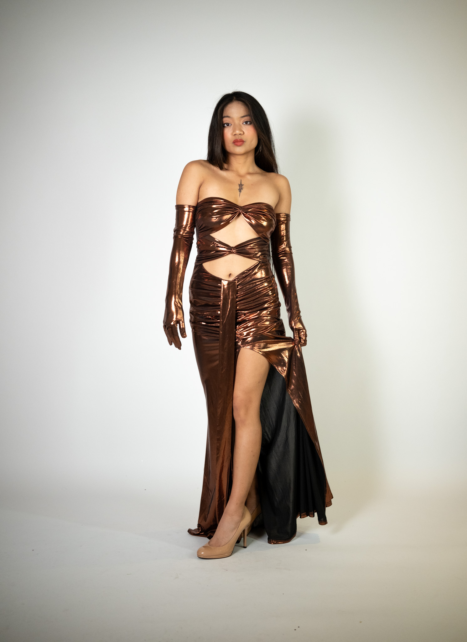 Alt text: "Bronze brown metallic strapless gown called Padame, featuring cutouts on the chest, ruching details, a high slit, and matching gloves, perfect for special occasions."