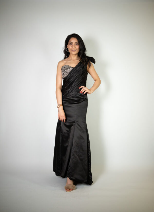 Alt text: "Black satin one-shoulder gown called Saar, featuring peek-a-boo rhinestone detailing on the breast, perfect for special occasions."