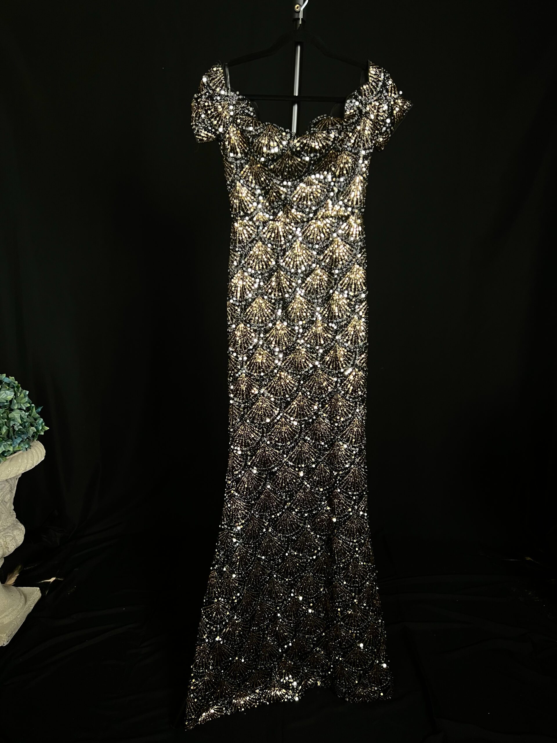 Introducing the Azalea Gown, a stunning black sequin dress adorned with silver and gold sequins that dazzle brilliantly under the lights. This elegant off-the-shoulder dress features a serrated neckline, adding a unique and sophisticated touch to its silhouette. Perfect for formal events, galas, and glamorous evenings out, the Azalea Gown ensures you stand out with elegance and style. Shine with confidence and grace in the captivating Azalea Gown. --- Alt text: "Black sequin dress called Azalea, with silver and gold sequins, off-the-shoulder design, and a serrated neckline, perfect for special occasions."